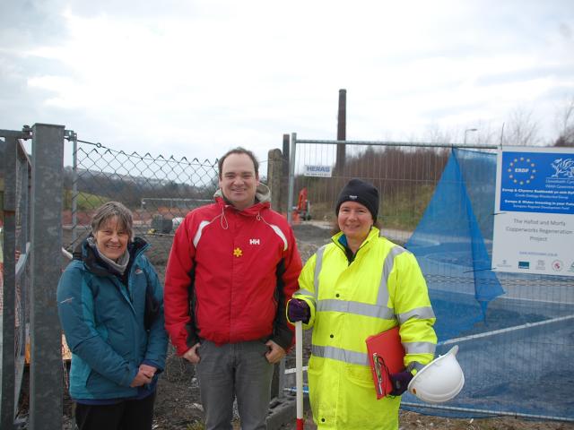 Three people standing by a chainlink fence with a sign saying 'The Hafod and Morfa Cooperworks Regeneration Project'