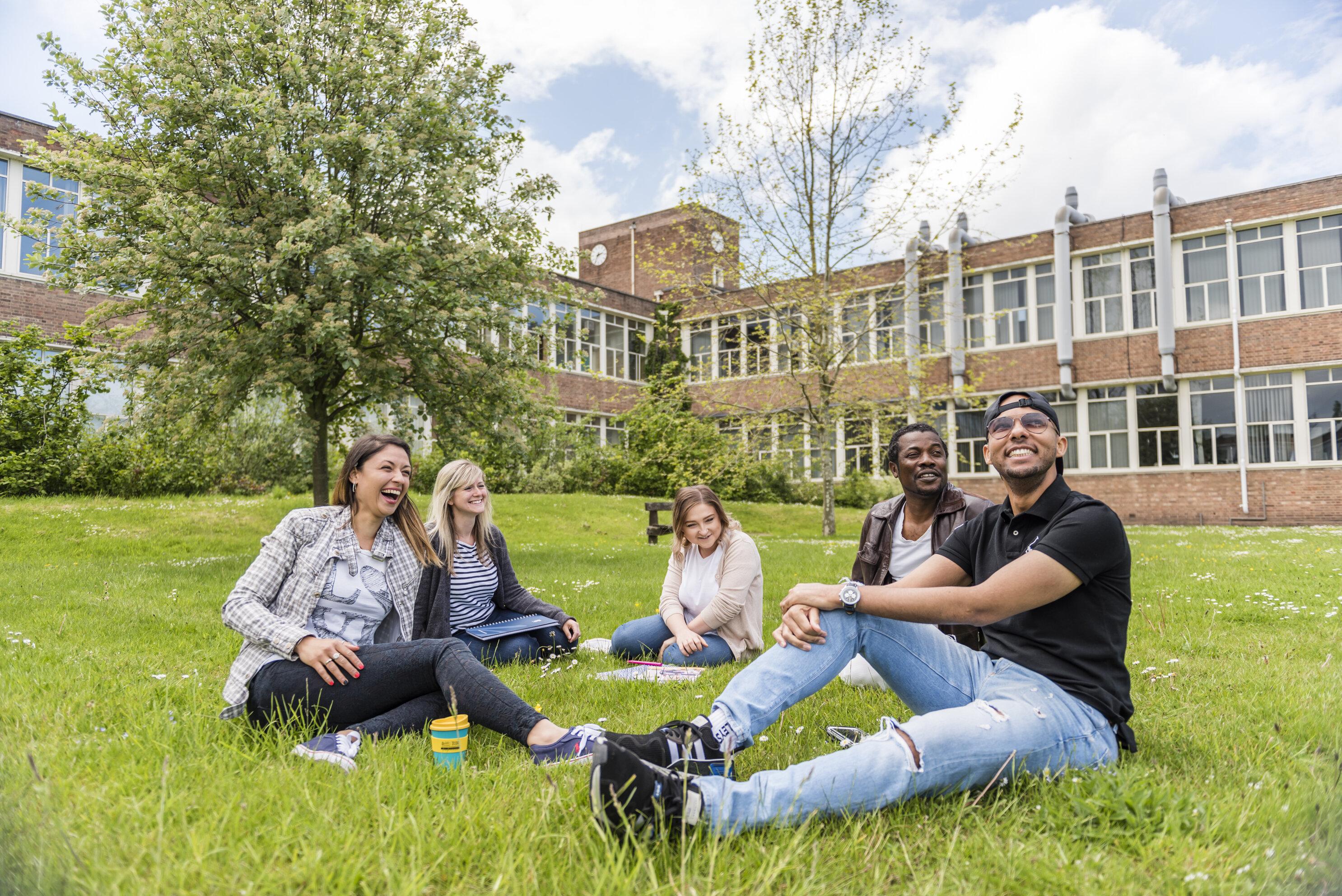 Three female and two male students sitting on a lawn outside a university building
