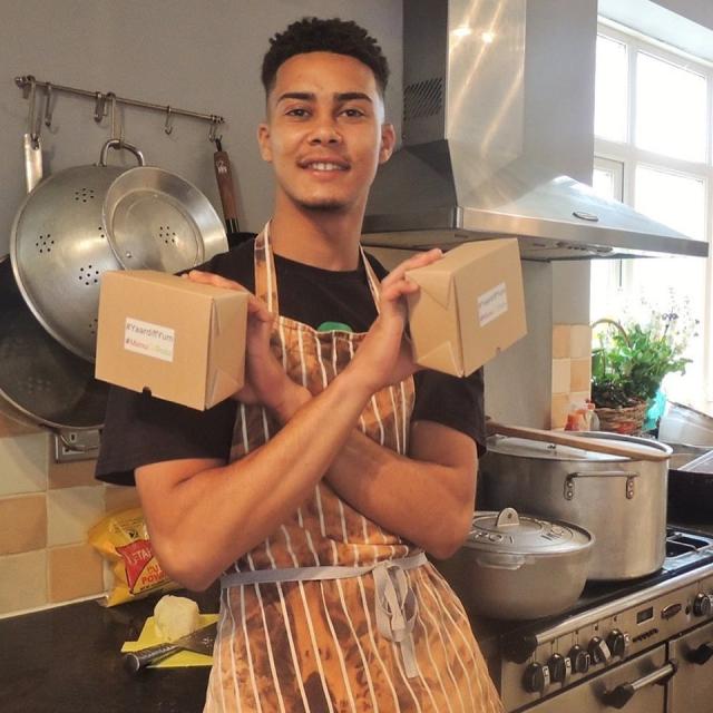 Young man standing in a kitchen, wearing an apron and holding two boxes