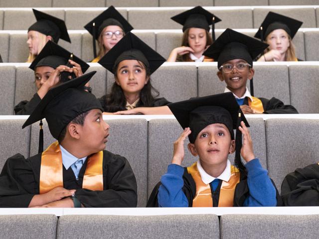 Young children in a lecture theatre wearing graduation caps and gowns